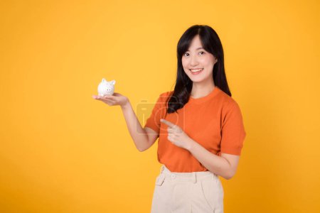 Photo for Smiling Asian woman 30s holding piggy bank, pointing to it, on vibrant yellow background. Wealth and saving concept. - Royalty Free Image