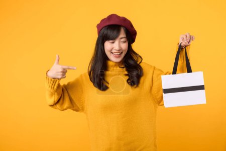 Téléchargez les photos : Jeune femme asiatique trentaine, tenant un sac en papier shopping, pointant son doigt avec une expression joyeuse, portant un pull jaune et un béret rouge sur un fond jaune. - en image libre de droit