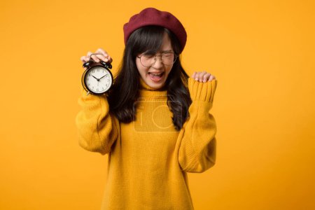 Téléchargez les photos : Une jeune femme asiatique au sourire heureux, portant un pull jaune et un béret rouge, se tient en confiance à côté d'un réveil dans son bureau élégant. - en image libre de droit