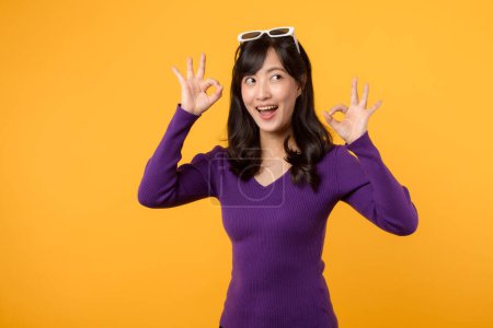 Foto de Bien, firma el gesto. Mujer asiática joven con camisa púrpura haciendo gesto de la mano OK con sonrisa segura feliz aislado en el fondo amarillo. De acuerdo, vale, concepto de gesto de mano aprobado. - Imagen libre de derechos