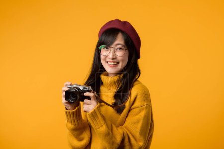 joven mujer asiática, una fotógrafa talentosa, captura recuerdos con su cámara, todo mientras exuda estilo en su suéter amarillo y boina roja contra un vibrante telón de fondo amarillo.