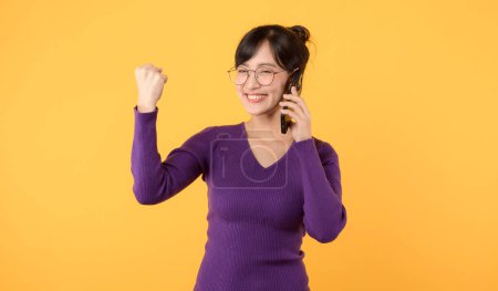 Téléchargez les photos : Dame heureuse, portant une chemise violette et des lunettes, embrassant la technologie avec son smartphone pour la joie sociale et le succès des achats en ligne. Restez intelligent, restez connecté sur un fond jaune! - en image libre de droit