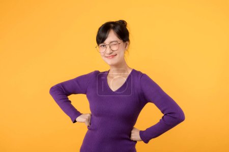 Photo for Portrait young asian woman wearing purple shirt and eyeglasses expression cheerful smile while hand holding hip isolated on yellow studio background. Attractive female model posing confident. - Royalty Free Image