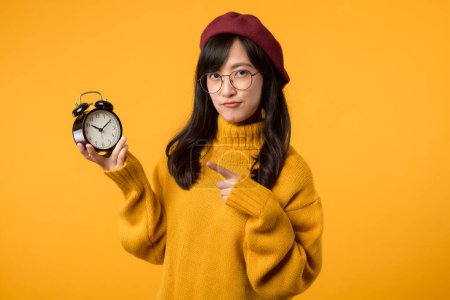 Téléchargez les photos : Une jeune asiatique ponctuelle, portant un pull jaune et un béret rouge, pointe son réveil, s'assurant qu'elle est toujours à l'heure, sur un fond jaune vif. - en image libre de droit