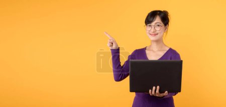 Téléchargez les photos : Une femme confiante, portant une chemise violette et des lunettes, tient un ordinateur portable et pointe du doigt pour libérer de l'espace sur un fond jaune, symbolisant son rôle de professionnelle numérique. - en image libre de droit