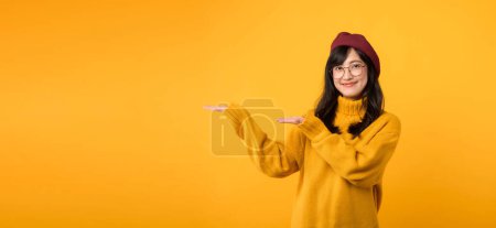 Téléchargez les photos : Magasinez avec style comme femme asiatique joyeuse trentaine dans un pull jaune, béret rouge et lunettes de vue pointe vendredi traite sur un fond jaune vibrant. - en image libre de droit