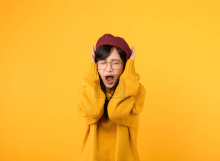Téléchargez les photos : Sensation de stress sonore. femme frustrée, dans un pull jaune et béret rouge, couvre ses oreilles sur un fond jaune. - en image libre de droit