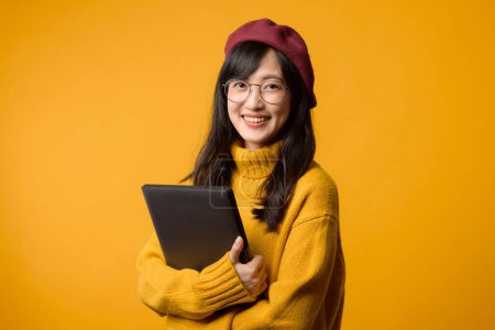 Foto de Destacando en suéter amarillo y boina roja, una joven asiática trabaja en su portátil sobre un alegre fondo amarillo. - Imagen libre de derechos