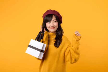 Foto de Sorprendido joven mujer asiática de 30 años, sosteniendo una bolsa de papel de compras, con elegancia en un suéter amarillo y boina roja sobre un fondo amarillo. - Imagen libre de derechos