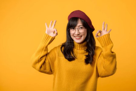 Jeune femme au béret rouge et pull jaune, faisant joyeusement le geste correct dans un environnement studio élégant.