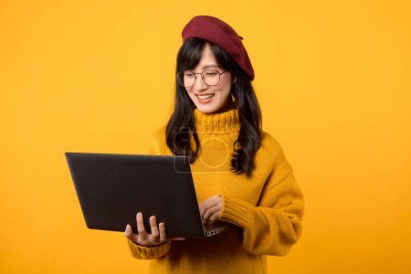 Foto de Retrato joven asiática estudiante vestida con suéter amarillo, boina roja y anteojos sonrisa feliz sosteniendo portátil aislado sobre fondo amarillo. concepto de educación - Imagen libre de derechos