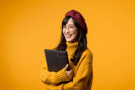 Téléchargez les photos : Sur fond jaune ensoleillé, une jeune femme asiatique utilise avec confiance son ordinateur portable tout en portant un pull jaune et un béret rouge. - en image libre de droit