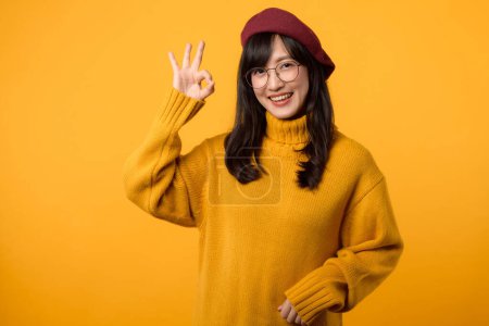 Foto de Mujer confiada en suéter amarillo, boina roja y gafas graduadas, haciendo el gesto correcto sobre un fondo amarillo vibrante. - Imagen libre de derechos