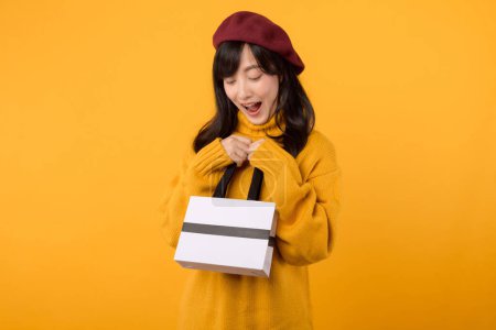 Téléchargez les photos : Jeune femme asiatique trentaine, portant un sac en papier shopping, portant un pull jaune et un béret rouge, profitant d'une séance de thérapie au détail sur fond jaune. - en image libre de droit