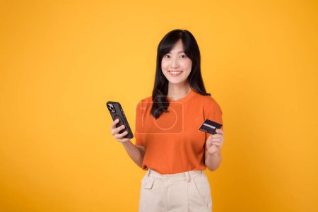 Photo for Cheerful young Asian woman in her 30s in orange shirt, using smartphone and holding credit card on yellow background. Effortless online shopping. - Royalty Free Image