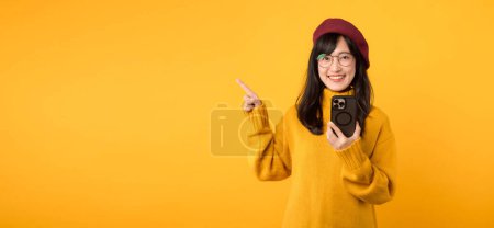 Foto de Retrato joven hermosa mujer asiática feliz sonrisa con camisa amarilla y sombrero rojo señalando gesto de dedo para liberar espacio de copia y el uso de teléfono inteligente contra el fondo amarillo. nueva aplicación - Imagen libre de derechos