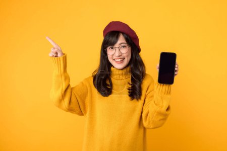 Foto de Comercialización digital delicia. Mujer joven, en alegre entorno amarillo, apunta a espacio de copia gratuita en su teléfono inteligente. - Imagen libre de derechos