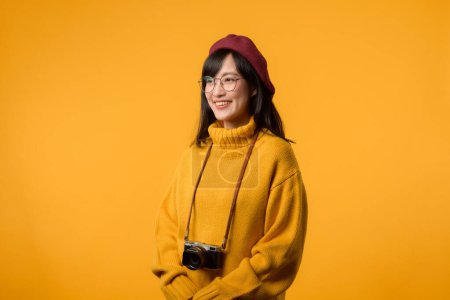 Con su cámara vintage en la mano, la joven asiática, vestida con un suéter amarillo y una boina roja, persigue su pasión por la fotografía sobre un vibrante fondo amarillo.