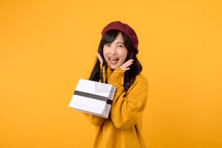 Téléchargez les photos : Surprise jeune femme asiatique trentaine, tenant un sac en papier shopping, mettant en valeur son sens de la mode avec un pull jaune et béret rouge sur un fond jaune. - en image libre de droit