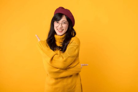 Foto de Mujer de moda, vestida con un suéter amarillo, boina roja y gafas graduadas, expresando su creatividad señalando con el dedo el espacio libre para copiar sobre un vibrante fondo amarillo. - Imagen libre de derechos