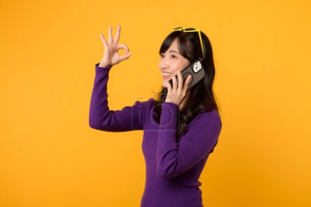 Foto de Irradiar alegría con la joven mujer asiática en sus 30 años, con una camisa púrpura, mostrando gesto de señal bien mientras se utiliza el teléfono inteligente sobre fondo amarillo. - Imagen libre de derechos