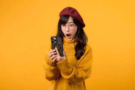 Foto de Una mujer joven, con la cara sorprendida, con una boina roja y un suéter amarillo, utiliza su teléfono inteligente sobre un fondo amarillo vibrante. - Imagen libre de derechos
