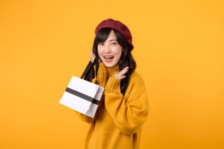 Téléchargez les photos : Jeune femme asiatique trentaine, avec une expression de surprise, portant un sac en papier shopping, profitant d'une séance de thérapie au détail dans un pull jaune et béret rouge sur un fond jaune. - en image libre de droit