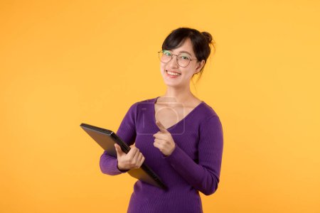 Téléchargez les photos : Jeune femme confiante, portant une chemise violette et des lunettes, tient un ordinateur portable sur un fond jaune vif, symbolisant le succès et le professionnalisme dans un portrait en studio. - en image libre de droit