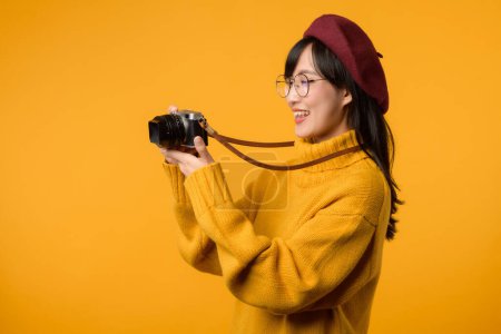 Foto de Con su cámara de confianza en la mano, la joven asiática explora el mundo, encarnando el espíritu de un fotógrafo en su elegante suéter amarillo y boina roja. - Imagen libre de derechos