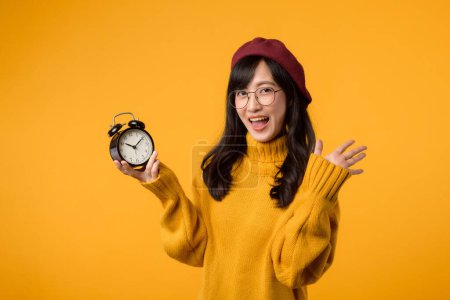 Photo for Close up of a vibrant yellow alarm clock held by a happy young Asian woman in a stylish yellow sweater and red beret, emphasizing the importance of time management. - Royalty Free Image