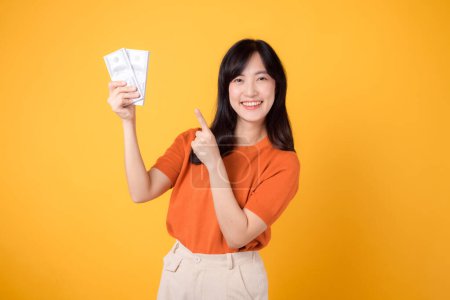Photo for Dynamic young Asian woman 30s, pointing finger to cash money dollars, standing on vibrant yellow backdrop. - Royalty Free Image