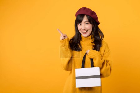 Téléchargez les photos : Jeune femme asiatique trentaine, tenant un sac en papier shopping, pointant du doigt, vêtue d'un pull jaune et d'un béret rouge sur un fond jaune. - en image libre de droit