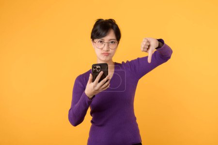 Téléchargez les photos : Jeune femme asiatique portant une chemise violette et des lunettes avec un visage malheureux tout en utilisant smartphone et pouce vers le bas mauvais signe geste de la main isolé sur fond jaune. - en image libre de droit