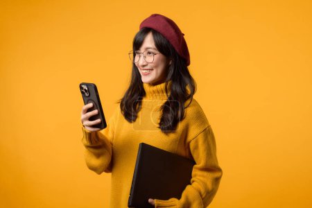 Téléchargez les photos : Sur un fond jaune ensoleillé, la jeune femme asiatique équilibre adepte son ordinateur portable et son smartphone, revêtant un pull jaune et un béret rouge. - en image libre de droit