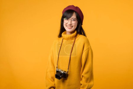 Foto de Una joven asiática, entusiasta de la cámara, con un suéter amarillo y una boina roja, captura recuerdos con su cámara vintage sobre un vibrante fondo amarillo. - Imagen libre de derechos