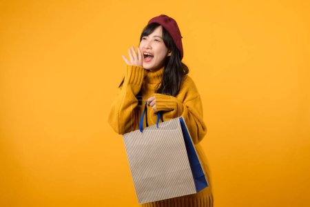 Téléchargez les photos : Jeune femme asiatique trentaine, vêtue d'un pull jaune et d'un béret rouge, criant de libérer de l'espace avec un sac en papier commercial sur fond jaune vif. - en image libre de droit