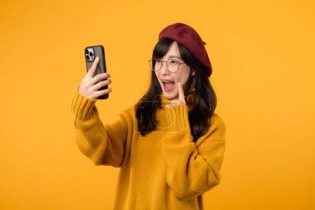 Téléchargez les photos : Une jeune femme joyeuse portant un béret rouge et un pull jaune prend un selfie avec son smartphone sur un fond jaune vibrant. - en image libre de droit
