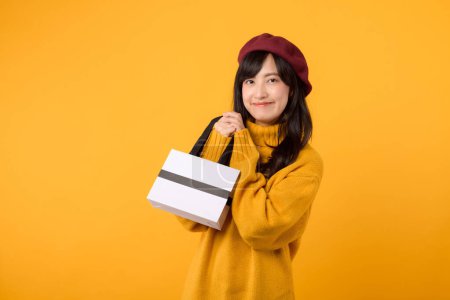 Photo for Young Asian woman 30s, with a shopping paper bag, showcasing her style in a yellow sweater and red beret against a yellow background. - Royalty Free Image