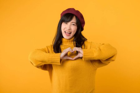 Téléchargez les photos : Jeune femme asiatique trentenaire, vêtue d'un pull jaune et d'un béret rouge, exprime son amour d'un geste de main de cœur sur un fond jaune. - en image libre de droit