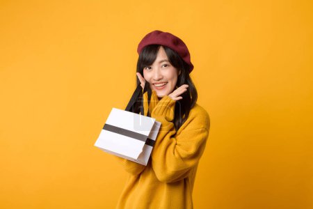 Téléchargez les photos : Jeune femme asiatique trentaine, avec une expression surprise, portant un sac en papier shopping, revêtu d'un pull jaune et d'un béret rouge sur un fond jaune. - en image libre de droit