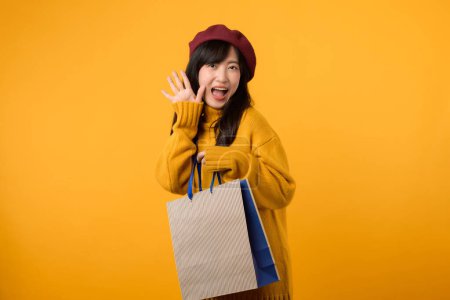 Photo for Young Asian woman 30s, wearing a yellow sweater and red beret, celebrates great deals while shopping against a vibrant yellow background. - Royalty Free Image