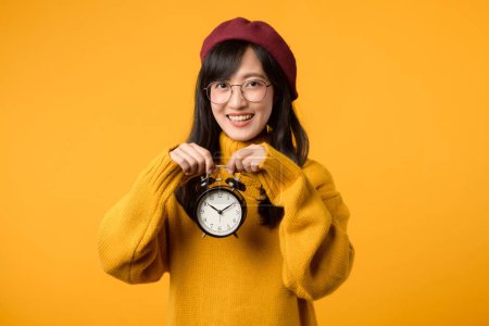 Close up of vibrant yellow alarm clock held by a young Asian woman, exuding happiness in her stylish yellow sweater and red beret.