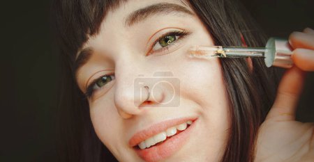 Téléchargez les photos : Une jolie brune au nez percé applique du sérum sur son visage - en image libre de droit