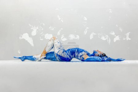 junge sexy Frau in Unterwäsche, Sportbekleidung, künstlerisch abstrakt mit weißer und blauer Farbe bemalt, auf dem Boden im Künstleratelier liegend, die Wand mit weißen Körperabdrücken, Kopierraum.