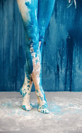 Blick von hinten auf schöne nackte Beine und Füße einer sexy jungen nackten Frau mit blauer und weißer Farbe auf dem Atelierboden des Künstlers, Kopierraum.