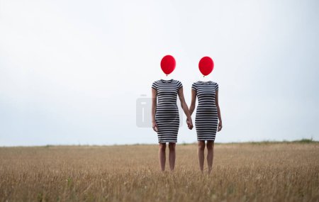 Photo for Two sexy young women, in black and white striped dress, friends, holding hands, heads replaced by red balloons stand hand in hand on harvested stubble field, copy space - Royalty Free Image