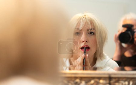 Foto de Mujer rubia se para en el espejo del baño y se aplica maquillaje, pinta los contornos de sus labios con un delineador de labios mientras es fotografiado por el fotógrafo, espacio para copiar - Imagen libre de derechos