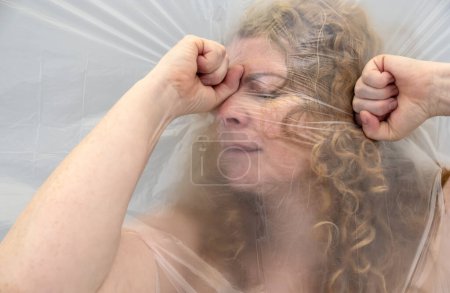 Téléchargez les photos : Image floue de l'art conceptuel d'une femme mûre saisissant sa tête avec ses mains d'un mal de tête, la position du visage derrière une feuille de film plastique transparent, représente la douleur terne, l'espace de copie - en image libre de droit
