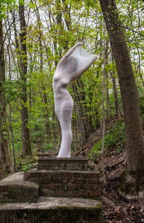 Foto de Sexy mujer adulta elegante de pie como estatua viviente, su cuerpo envuelto en piel larga vestido blanco apretado, destacando las formas del cuerpo, en el bosque de primavera verde en un pedestal de ladrillo, obra de arte, espacio de copia - Imagen libre de derechos