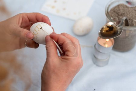 Las manos femeninas pintan un huevo de Pascua blanco según la tradición sorbia con una pluma de ganso recortada y cera de abeja y vela derretida, espacio para copiar